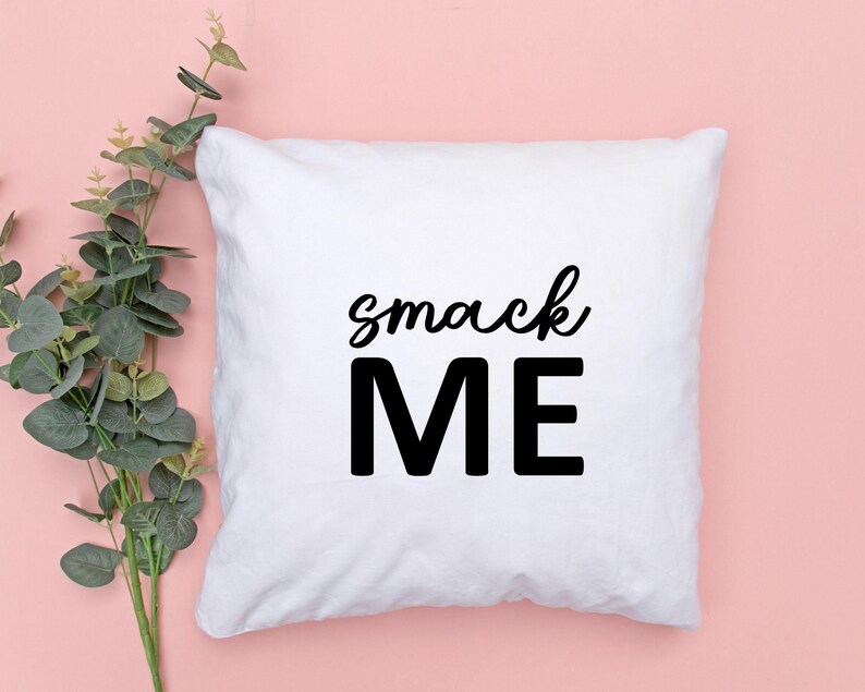 May include: White square pillow with the words "smack ME" printed in black. The word "smack" is in a cursive font, and "ME" is in a bold, block font. The pillow is on a pink background with green foliage.