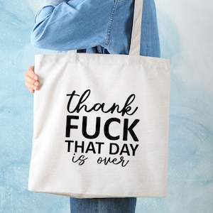 May include: A white tote bag with black text that reads "thank FUCK THAT DAY is over". The bag has two handles and is held by a person wearing a denim shirt. The background is a light blue.