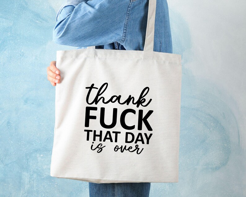 May include: A white tote bag with black text that reads "thank FUCK THAT DAY is over". The bag has two handles and is held by a person wearing a denim shirt. The background is a light blue.