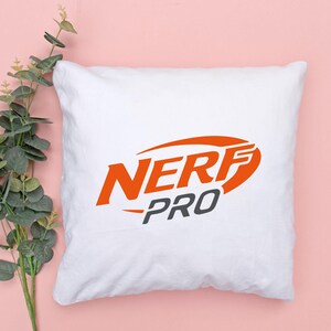 May include: White square pillow with the orange and gray "NERF PRO" logo. The pillow is on a pink background with green and gray leaves.