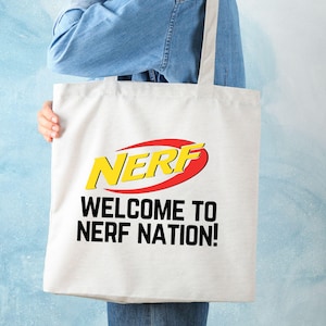 May include: White tote bag with the word "NERF" in yellow and red, with the text "WELCOME TO NERF NATION!" in black. The bag is held by a person wearing a denim shirt.