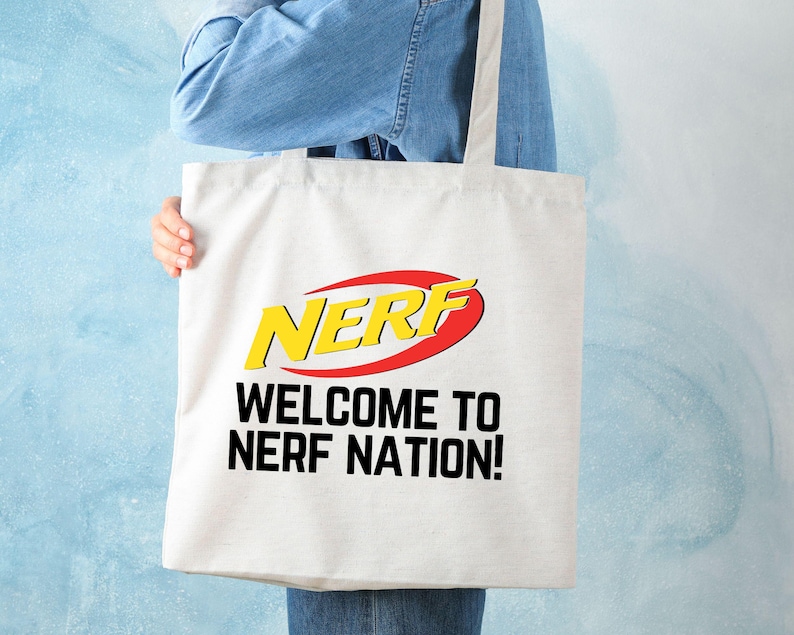 May include: White tote bag with the word "NERF" in yellow and red, with the text "WELCOME TO NERF NATION!" in black. The bag is held by a person wearing a denim shirt.