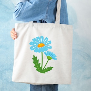 May include: A white tote bag featuring a blue daisy design with a yellow center and green leaves. The tote bag is held by a person wearing a denim shirt and jeans. The bag is made of a sturdy material, perfect for carrying groceries or other items.