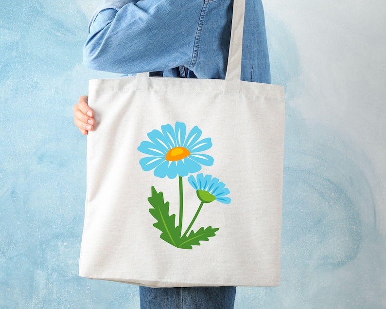 May include: A white tote bag featuring a blue daisy design with a yellow center and green leaves. The tote bag is held by a person wearing a denim shirt and jeans. The bag is made of a sturdy material, perfect for carrying groceries or other items.