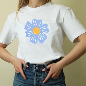 May include: White t-shirt featuring a large, stylized blue flower with a yellow center. The flower design is centered on the front of the shirt. The shirt has short sleeves and a crew neck.