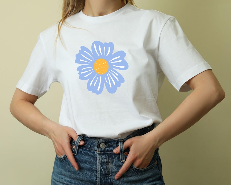 May include: White t-shirt featuring a large, stylized blue flower with a yellow center. The flower design is centered on the front of the shirt. The shirt has short sleeves and a crew neck.