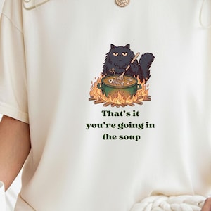 May include: An off-white short-sleeve t-shirt featuring a graphic of a black cat stirring a green pot over a campfire. Below the illustration, the text reads: That's it you're going in the soup. The shirt has a crew neckline and a relaxed fit.