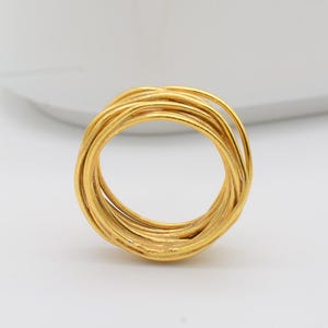 May include: A gold-colored ring with a woven design, featuring multiple intertwined bands. The ring is set against a white background, with a white, rectangular dish in the background.