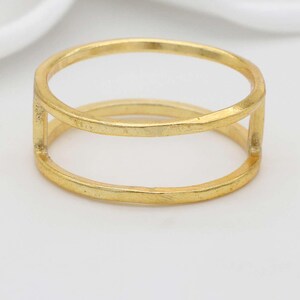 Puede incluir: Un anillo dorado con un diseño de doble banda. El anillo tiene forma rectangular con dos bandas paralelas conectadas por barras verticales. El anillo está sobre un fondo blanco.