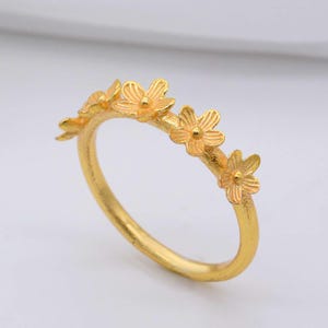 May include: A gold-colored ring featuring a delicate floral design. The ring has a band adorned with small, detailed flowers. The flowers are evenly spaced around the ring, creating a continuous floral pattern. The ring is set against a white background.