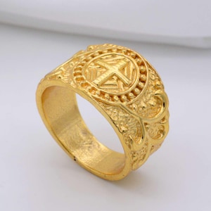 Peut inclure: Une bague dorée avec un motif de croix au centre. La bague a une bande ornée et texturée et un emblème circulaire avec une croix. La bague est posée sur un fond blanc.