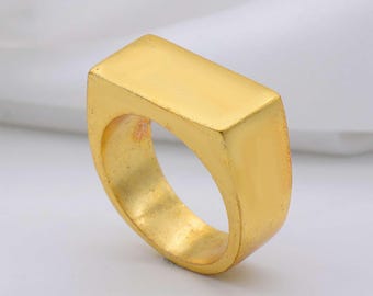Könnte beinhalten: Ein goldfarbener Ring mit rechteckiger Oberseite. Der Ring hat eine glatte, polierte Oberfläche und einen abgerundeten Ring. Der Ring ist ein solides, geometrisches Design, geeignet für den Alltag oder besondere Anlässe.