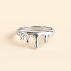 May include: A silver ring with a unique design. The ring features a band with a series of stylized drips cascading down the front. The ring is made of a shiny, metallic material and is set against a neutral background.