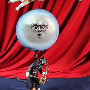 May include: A whimsical sculpture featuring a brown and white cow jumping over a moon with a surprised face wearing glasses. A black cat with white paws plays a violin on a stand below the moon. The sculpture is on a wooden base with three red stars.