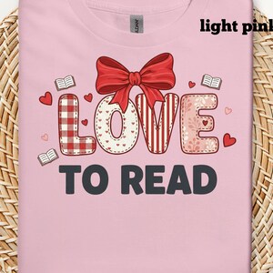 Op de afbeelding: Lichtroze T-shirt met de tekst "LOVE TO READ" in een decoratief ontwerp. De "L" heeft een rood-wit ruitpatroon, de "O" is wit met kleine hartjes, de "V" is rood-wit gestreept en de "E" is roze met bloemmotieven. Een rode strik staat boven het woord "LOVE".