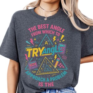 May include: Dark grey t-shirt with a colourful graphic design. The design features the text "THE BEST ANGLE FROM WHICH TO TRY ANGLE APPROACH A PROBLEM IS THE" with mathematical symbols and a calculator.
