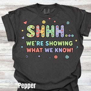 May include: Pepper-colored t-shirt with the text "SHHH... WE'RE SHOWING WHAT WE KNOW!" in colorful bubble letters. The design includes hearts, stars, smiley faces, and other playful graphics. A Comfort Colors brand tag is visible.