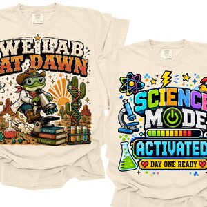 Könnte beinhalten: Zwei cremefarbene T-Shirts mit wissenschaftlichen Motiven. Das linke T-Shirt zeigt einen Frosch im Laborkittel mit dem Text "WE LAB AT DAWN" und Laborausrüstung. Das rechte T-Shirt zeigt den Text "SCIENCE MODE ACTIVATED" mit einer Rakete, einem Mikroskop und einem DNA-Strang.