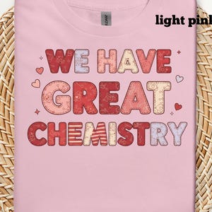 May include: Pale pink t-shirt featuring the text "WE HAVE GREAT CHEMISTRY" in a patchwork design. The words are in red, pink, and cream, with heart and star embellishments. The shirt is presented on a woven background.