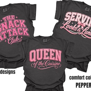 Op de afbeelding: Drie antracietgrijze t-shirts met roze grafische ontwerpen. Eén shirt heeft de tekst THE SNACK ATTACK CLUB, een ander SERVING Looks & Lunch met een roze strik, en het derde QUEEN of the Cuisine. De shirts zijn gelabeld als comfort colors pepper.