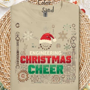 May include: Sand-colored t-shirt with a Christmas-themed design. The design includes a Santa hat, snowflakes, and the words "ENGINEERING CHRISTMAS CHEER" in red and green block letters. The shirt is displayed on a woven basket.