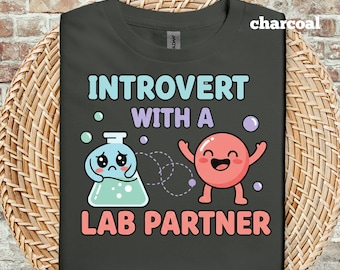 Op de afbeelding: Antracietkleurig T-shirt met de tekst "INTROVERT WITH A LAB PARTNER". Het ontwerp toont een trieste cartoonkolf en een vrolijke cartooncel. De tekst en afbeeldingen zijn in tinten blauw, roze en paars.