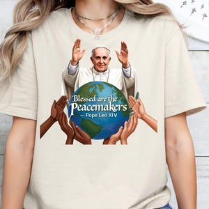 May include: A natural-colored t-shirt featuring a graphic of Pope Francis with his hands raised, holding a globe. The globe is surrounded by hands of various skin tones. Text on the shirt reads "Blessed are the Peacemakers - Pope Leo XIV."