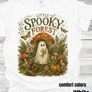 May include: A white t-shirt featuring a graphic print with the text "LITTLE SPOOKY FOREST" above a ghost standing under a large red mushroom. The design includes various forest elements like butterflies, smaller mushrooms, a pumpkin, and snails.