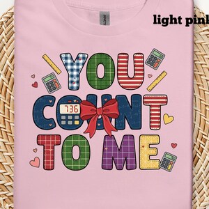 May include: Light pink t-shirt with the phrase "YOU COUNT TO ME" in colorful, patterned letters. The design includes calculators, rulers, hearts, and a red bow. The shirt is made of a soft material.