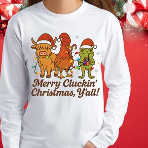 May include: White long-sleeve shirt featuring a festive graphic. The design includes a Highland cow, a chicken, and a frog, all wearing Santa hats and wrapped in Christmas lights. The text reads "Merry Cluckin' Christmas, Y'all!"