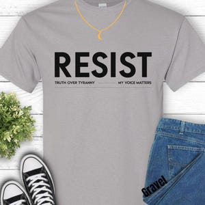 May include: A light gray t-shirt with the word "RESIST" in large black letters. Below the word are the phrases "TRUTH OVER TYRANNY" and "MY VOICE MATTERS". A gold necklace with a crescent moon pendant is visible.