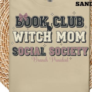 Może przedstawiać: Piaskowa koszulka z napisem "BOOK CLUB WITCH MOM SOCIAL SOCIETY" w stylu retro. Projekt zawiera sylwetkę czarnego kota i różową kokardę. Tekst "Branch President" jest również uwzględniony.