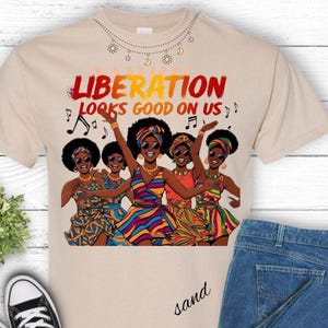 Może przedstawiać: T-shirt w kolorze piasku z grafiką przedstawiającą pięć kobiet z afro i okularami przeciwsłonecznymi, ubranych w kolorowe sukienki. Tekst "LIBERATION LOOKS GOOD ON US" jest wydrukowany nad ilustracją, z nutami. Koszulka jest połączona z niebieskimi dżinsami.