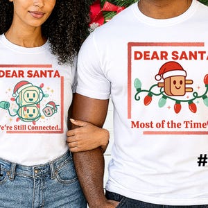 Op de afbeelding: Twee witte T-shirts met kerstthema's. Eén shirt heeft een stekker- en stopcontactontwerp met de tekst "Dear Santa, We're Still Connected." Het andere shirt heeft een stekkerontwerp met de tekst "Dear Santa, Most of the Time."