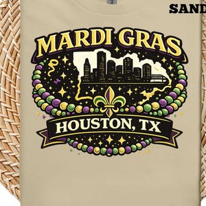 Könnte beinhalten: Sandfarbenes T-Shirt mit einem Mardi Gras-Design. Das Design zeigt die Worte "Mardi Gras" in Gelb und Lila, eine Silhouette der Skyline einer Stadt und den Text "Houston, TX". Das Design ist von grünen, lila und gelben Perlen umgeben.