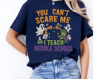 May include: Navy blue t-shirt with a Halloween-themed graphic. The design features the text "YOU CAN'T SCARE ME I TEACH MIDDLE SCHOOL" in orange, green, and purple. The graphic includes ghosts, a zombie, and a haunted house.