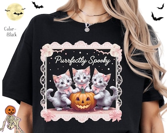 Puede incluir: Camiseta negra con un diseño temático de Halloween. El gráfico incluye tres gatitos grises con lazos rosas, una calabaza tallada y el texto "Purrfectly Spooky" dentro de un marco decorativo. También hay murciélagos y una luna llena.
