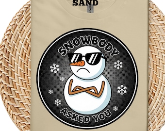 Pode incluir: Camiseta cor de areia com um gráfico de um boneco de neve usando óculos de sol e braços cruzados. O texto "SNOWBODY ASKED YOU" está em um design circular ao redor do boneco de neve, com flocos de neve. A palavra "SAND" está impressa no topo.