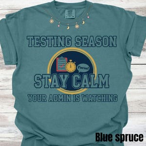 May include: Blue spruce t-shirt with the words "TESTING SEASON STAY CALM YOUR ADMIN IS WATCHING". The shirt features a circular graphic with a clipboard and stopwatch. The shirt also has a decorative neckline.