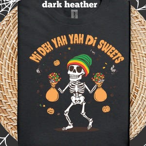Puede incluir: Camiseta de color gris oscuro con un diseño de esqueleto que lleva un gorro verde, amarillo y rojo. El esqueleto sostiene dos bolsas naranjas llenas de flores de colores. El texto en la camiseta dice "Mi Deh Yah Di Sweets".