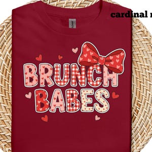 May include: A cardinal red t-shirt with the words "BRUNCH BABES" in a playful font. The design includes a red bow with heart patterns and small hearts scattered around the text. The shirt is displayed on a woven surface.