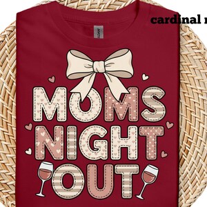 May include: Cardinal red t-shirt with the text "MOMS NIGHT OUT" in a decorative font, featuring a bow and wine glasses. The letters have a stitched design with heart accents. The shirt is displayed on a woven surface.