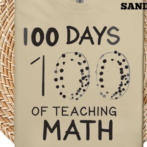 May include: A sand-coloured t-shirt featuring the text "100 DAYS OF TEACHING MATH" in black. The number "100" is stylized with dotted circles. The word "SAND" is printed in the upper right corner.