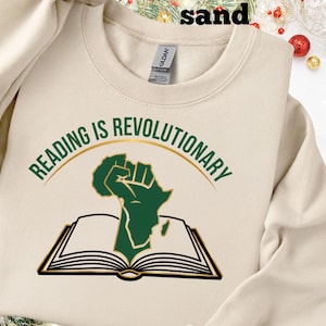 May include: Sand-coloured sweatshirt featuring the text "READING IS REVOLUTIONARY" in green above a green fist and open book graphic. The sweatshirt is a Gildan brand.
