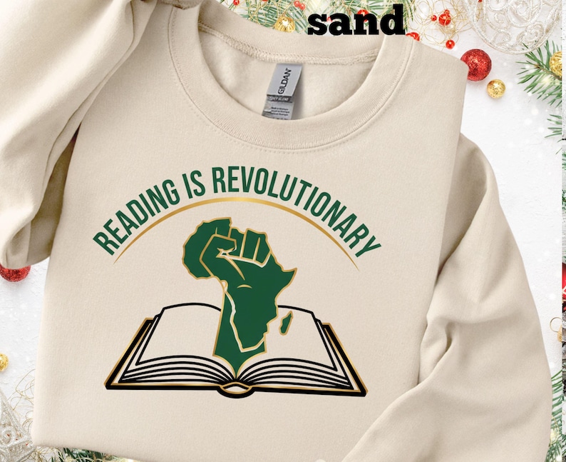 May include: Sand-coloured sweatshirt featuring the text "READING IS REVOLUTIONARY" in green above a green fist and open book graphic. The sweatshirt is a Gildan brand.