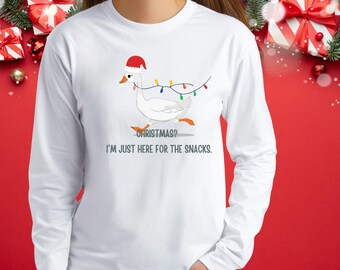 May include: White long-sleeve shirt featuring a cartoon goose wearing a Santa hat and Christmas lights. The text reads "Christmas? I'm just here for the snacks."