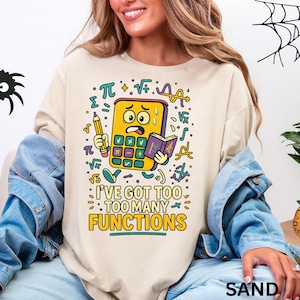 Puede incluir: Sudadera color arena con un gráfico humorístico de una calculadora estresada sosteniendo un lápiz y un libro, rodeada de símbolos matemáticos. El texto dice: "I'VE GOT TOO MANY FUNCTIONS".
