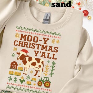 Peut inclure: Sweat-shirt couleur sable avec un motif festif. Le graphisme représente une vache portant un bonnet de Père Noël, une grange et le texte "MOO-Y CHRISTMAS Y'ALL" en marron. Le motif comprend des accents verts, rouges et dorés, créant un thème de vacances.