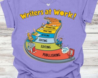 May include: Violet-colored t-shirt with the text "Writers at Work!" in yellow. The graphic depicts the writing process as a path with stages like prewriting, drafting, and publishing. A silver heart-shaped pendant hangs from the neckline.