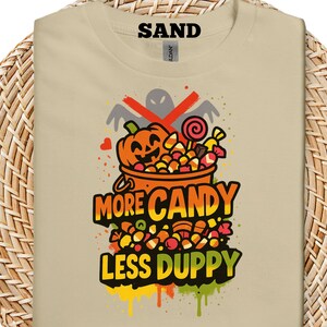 Puede incluir: Camiseta color arena con un diseño de Halloween. El diseño incluye una calabaza, un cubo lleno de caramelos y el texto "MORE CANDY LESS DUPPY". Un fantasma está tachado encima de la calabaza.